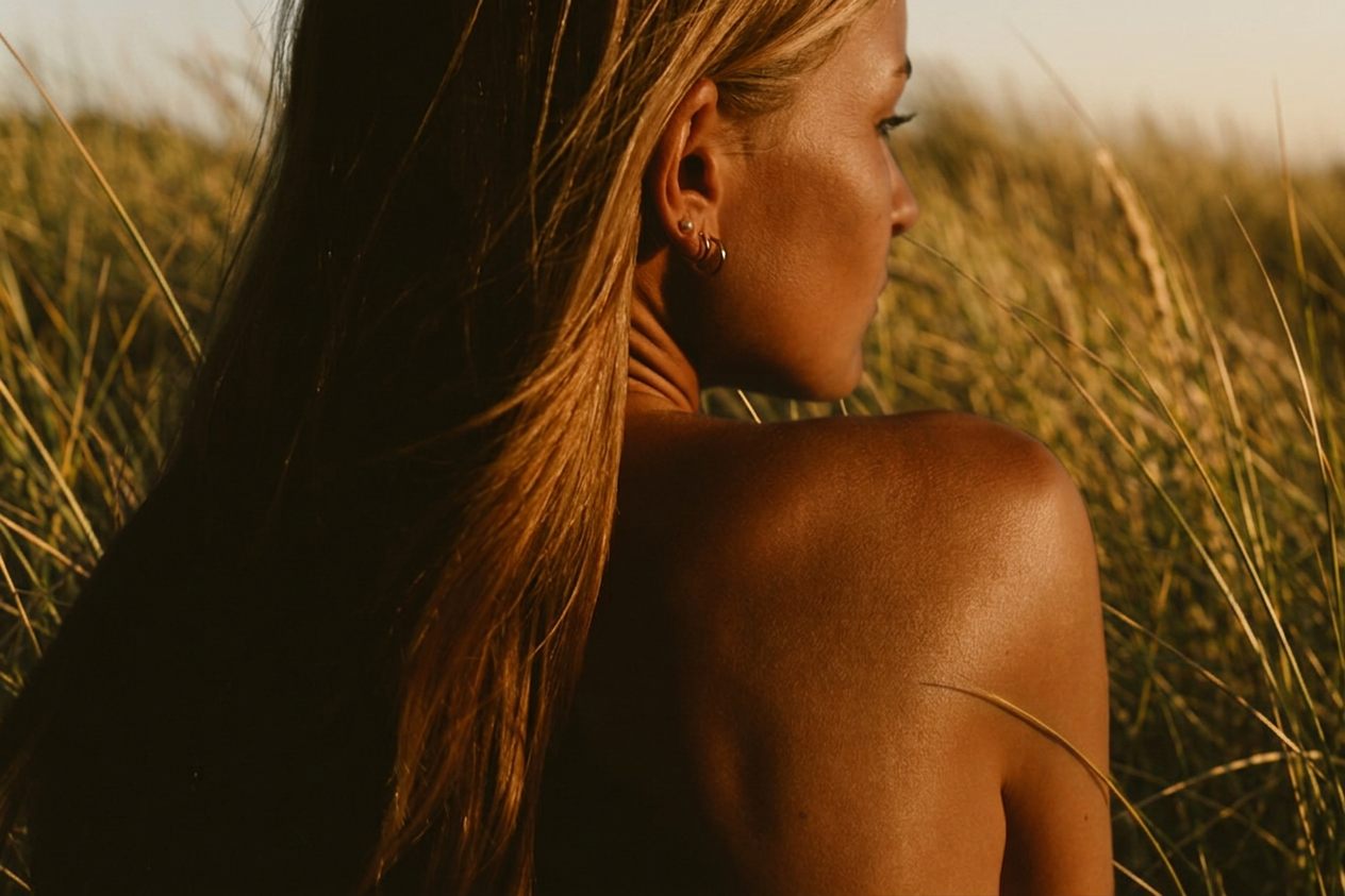 A woman with long brown hair and earrings looking into a field of tall grass during sunset.