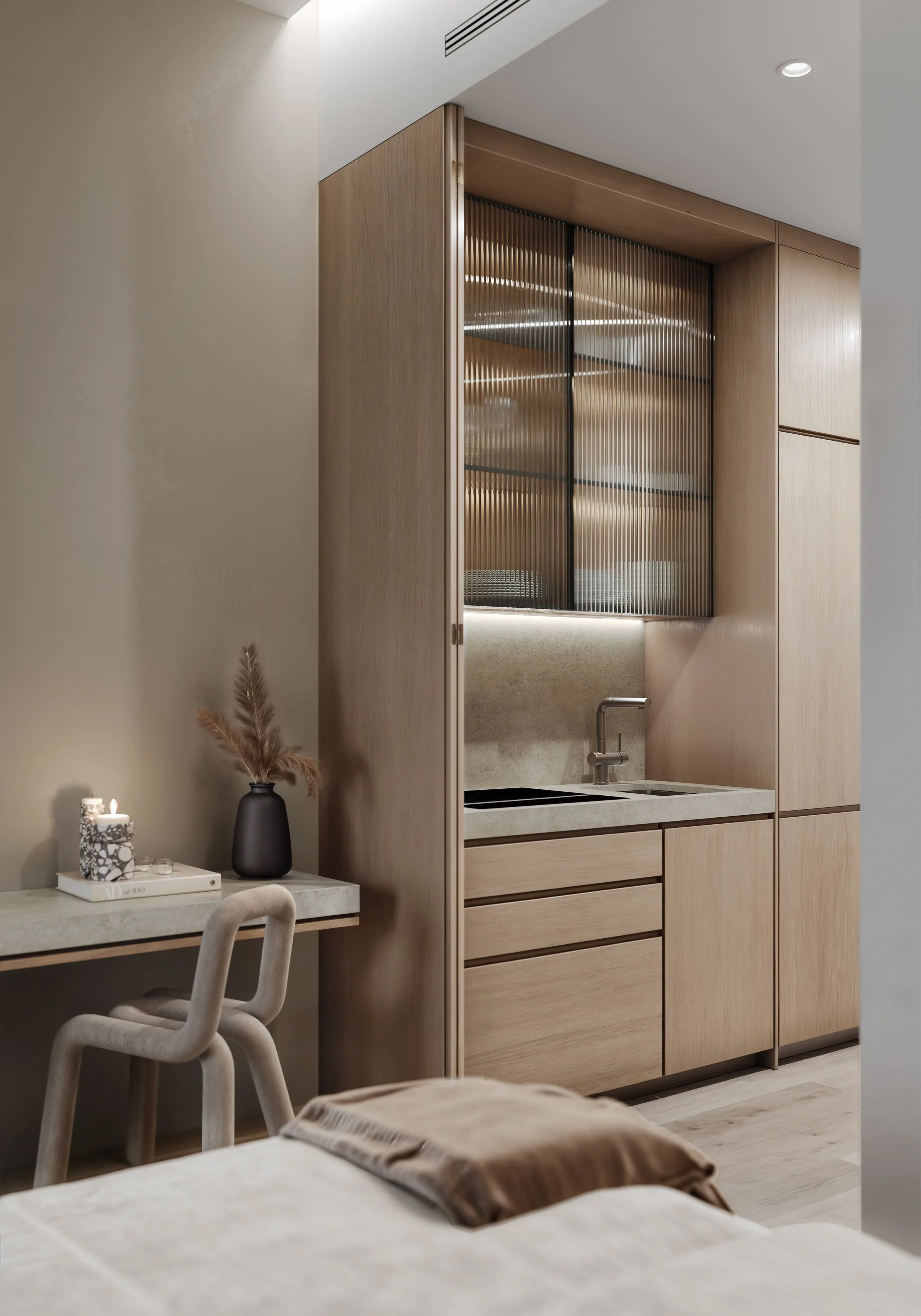Modern interior with a small kitchen area featuring wooden cabinetry, a sink, and a nest of black glass sliding doors, with a nearby table decorated with candles, a vase with dried plants, and books, and part of a bed with a blanket in the foreground.