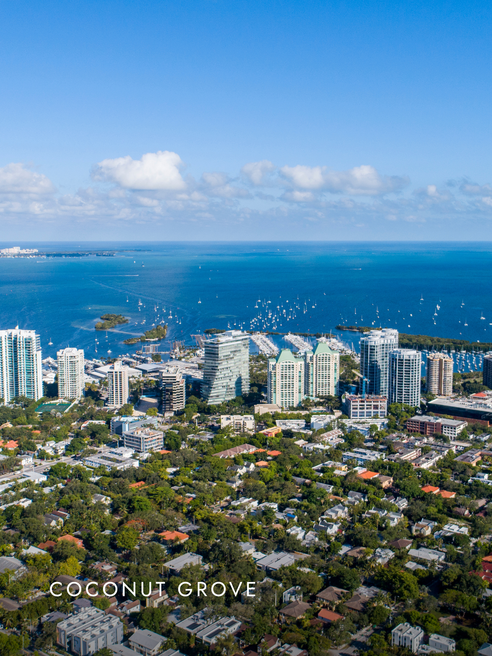 An aerial view of Coconut Grove, Miami, showing high-rise buildings, residential houses, a marina with boats, and the Atlantic Ocean in the background under a partly cloudy sky.