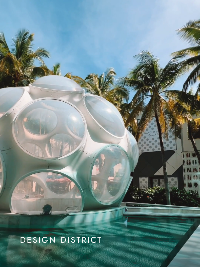A futuristic, spherical building with large glass windows in the Design District, surrounded by a pool and tall palm trees, under a clear blue sky.