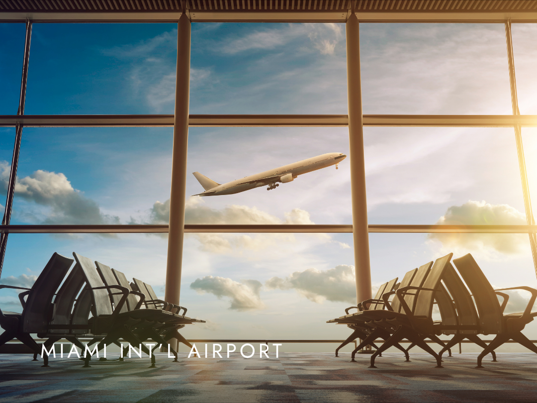 View of Miami International Airport terminal interior with empty chairs facing a large window showing an airplane taking off against a partly cloudy sky during sunset.
