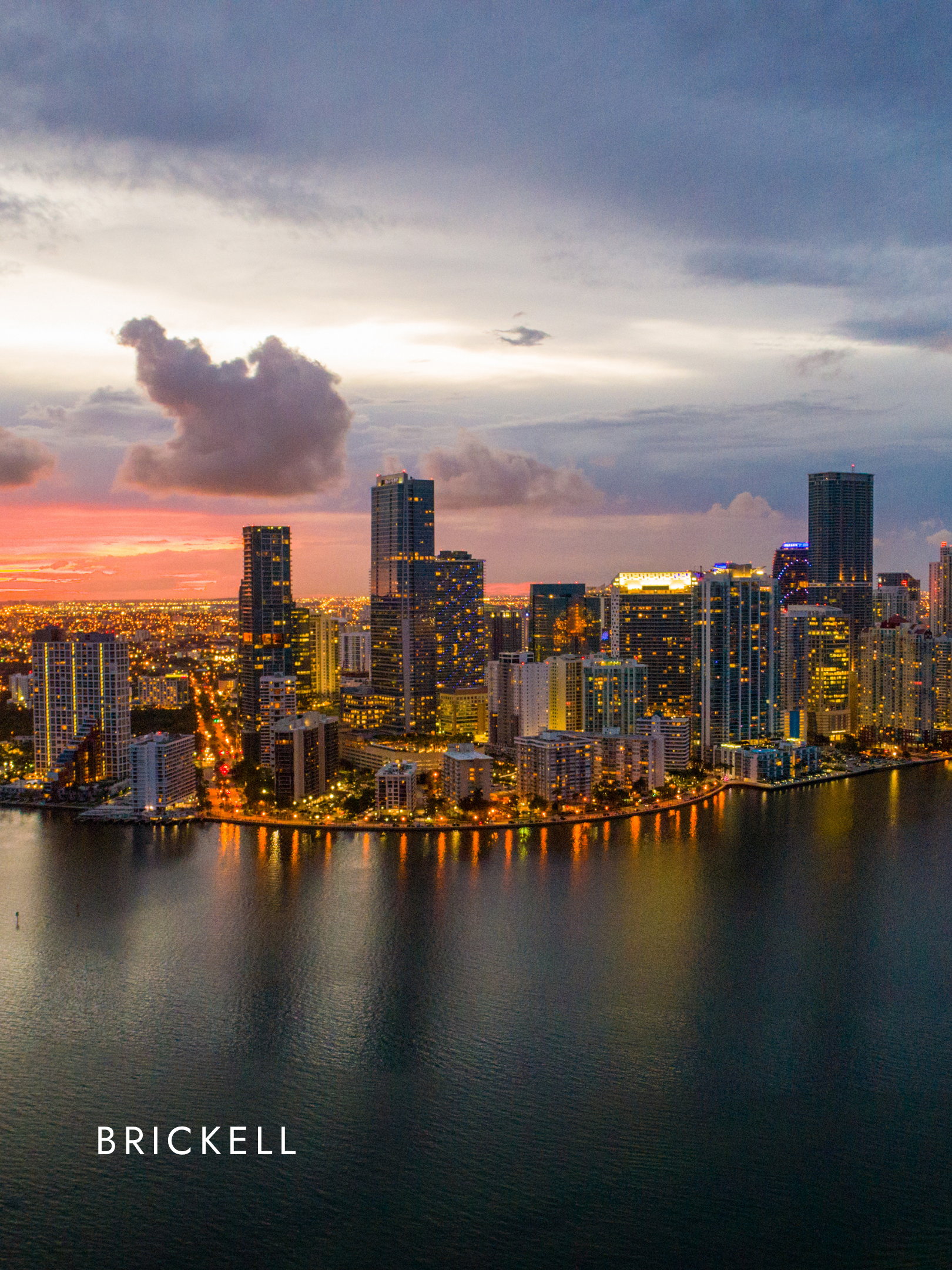 Brickell city skyline at sunset with tall buildings, clouds in the sky, and a body of water in the foreground, with the word 'BRICKELL' displayed at the bottom.