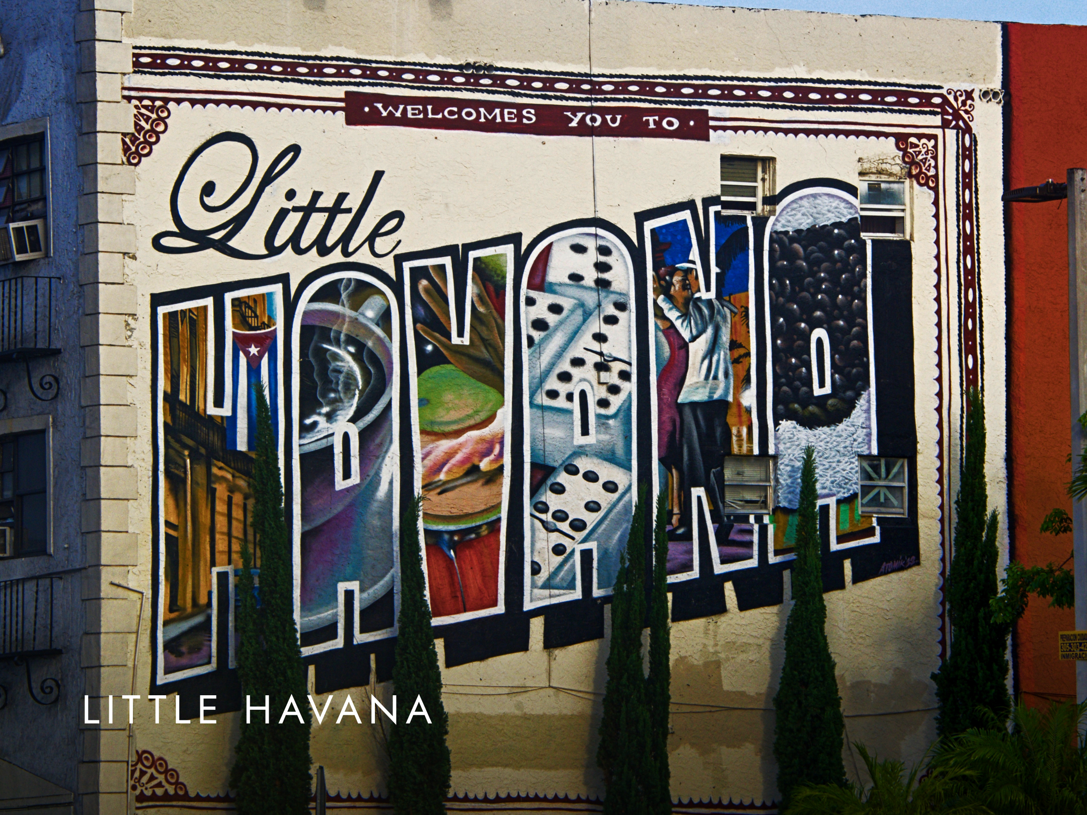 A colorful mural on a building wall that says 'Welcomes You to Little Havana' with various artistic elements, including images of dominoes, a woman, and vibrant patterns, surrounded by small trees at the base.