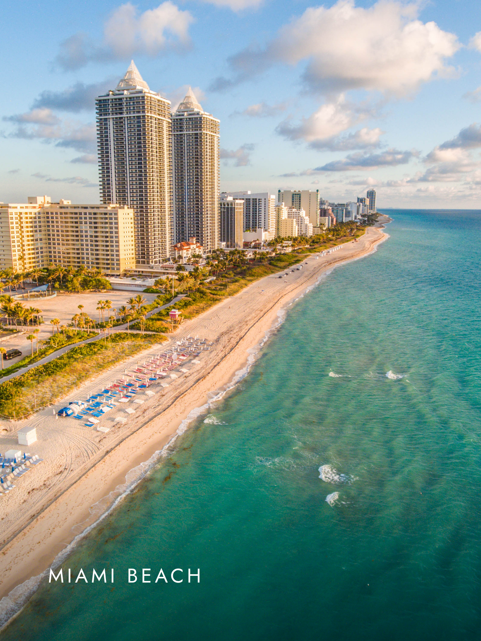 Aerial view of Miami Beach showing tall skyscrapers, sandy beach with colorful umbrellas, and the ocean with waves under a partly cloudy sky.