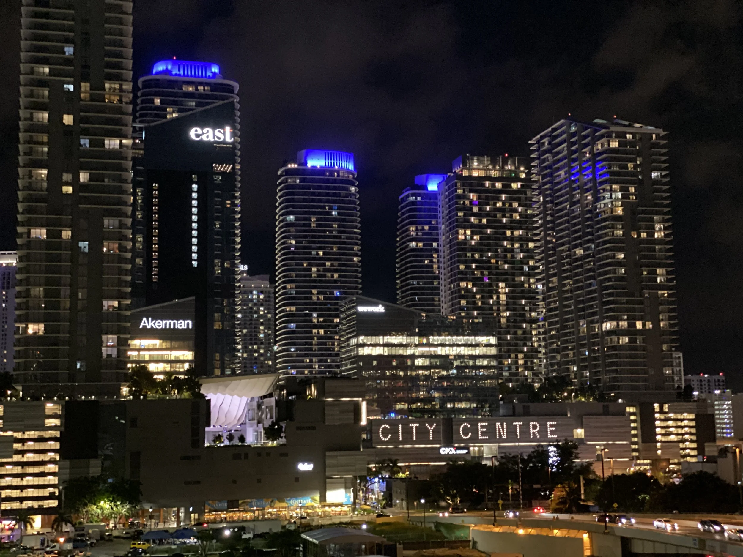 Nighttime cityscape of East Miami at Brickell City Centre with illuminated blue and white lights and a sign reading 'CITY CENTRE' at the lower part of the image.