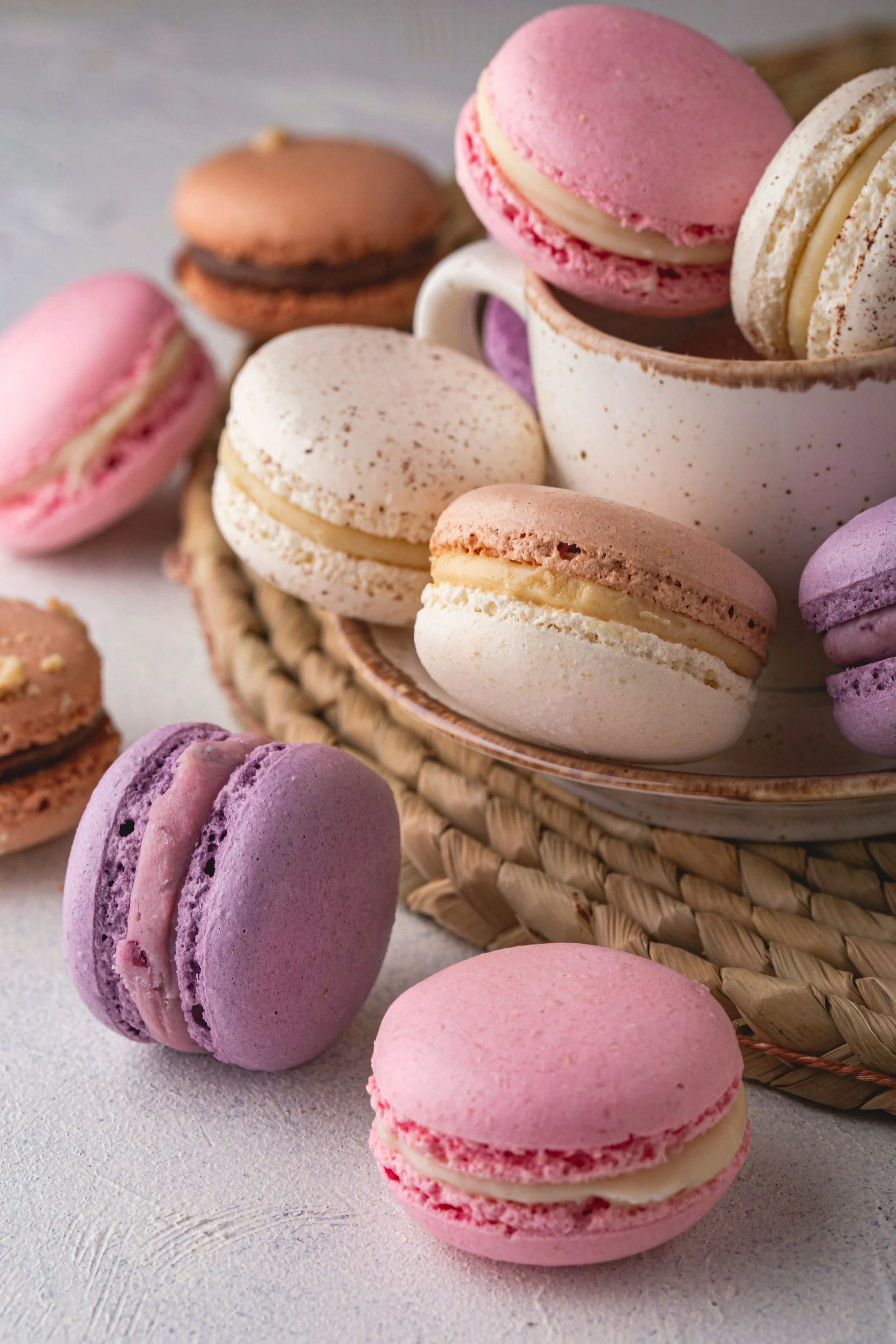 Assorted pastel-colored macarons in pink, purple, white, and brown, some inside a woven basket and others scattered on a light textured surface.