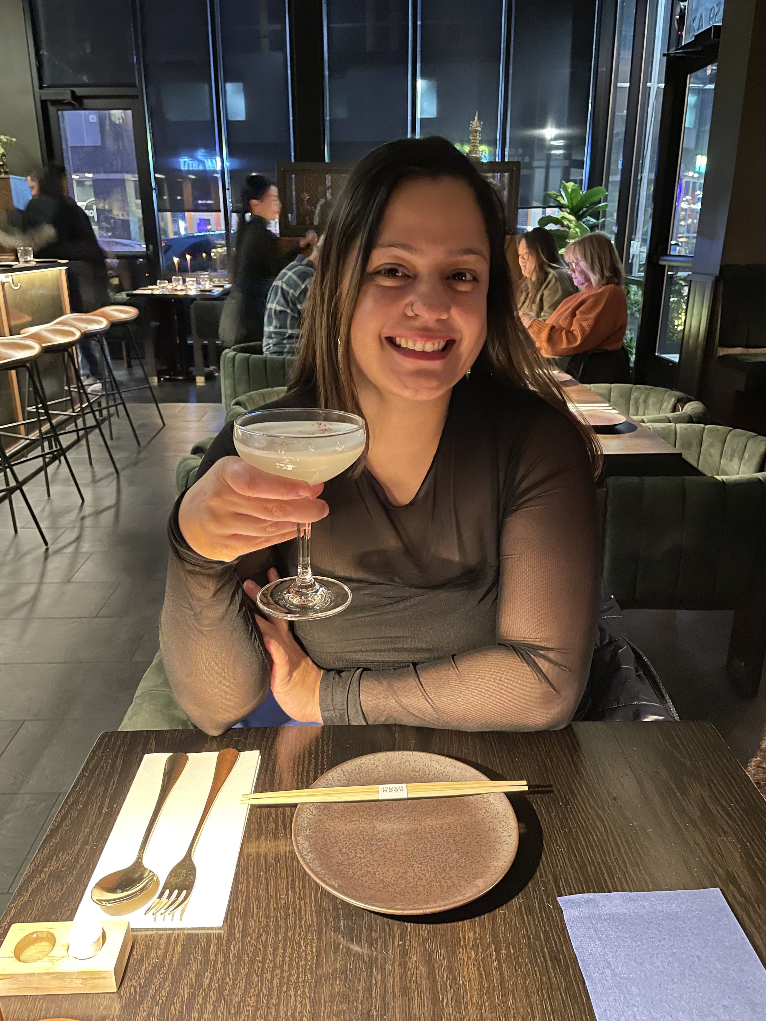 A woman sitting at a restaurant table holding a cocktail glass, smiling at the camera. The table has a set of cutlery on a white paper placemat, with a small empty plate and a napkin. The restaurant has a modern interior with large windows and other patrons in the background.