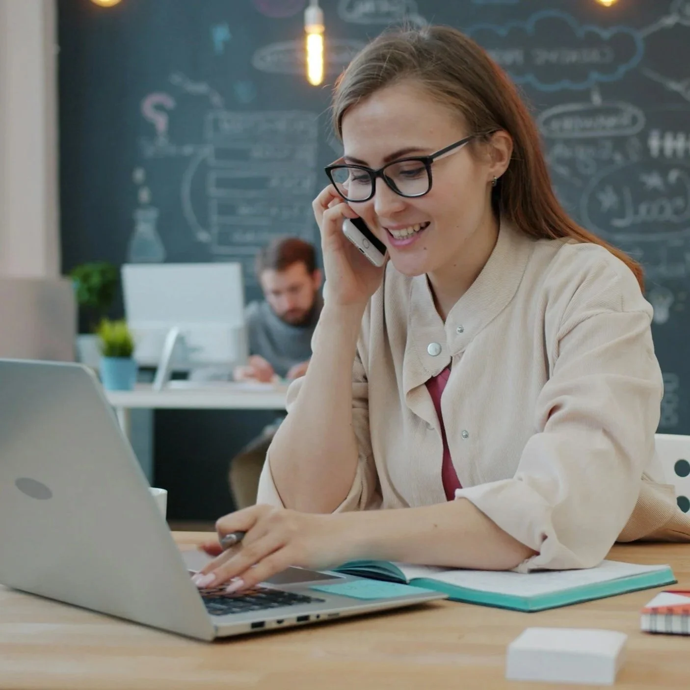 A woman wearing glasses sitting at a desk, talking on the phone, with a laptop and notebook in front of her. In the background, a man is working at another desk with a chalkboard covered in drawings and notes.