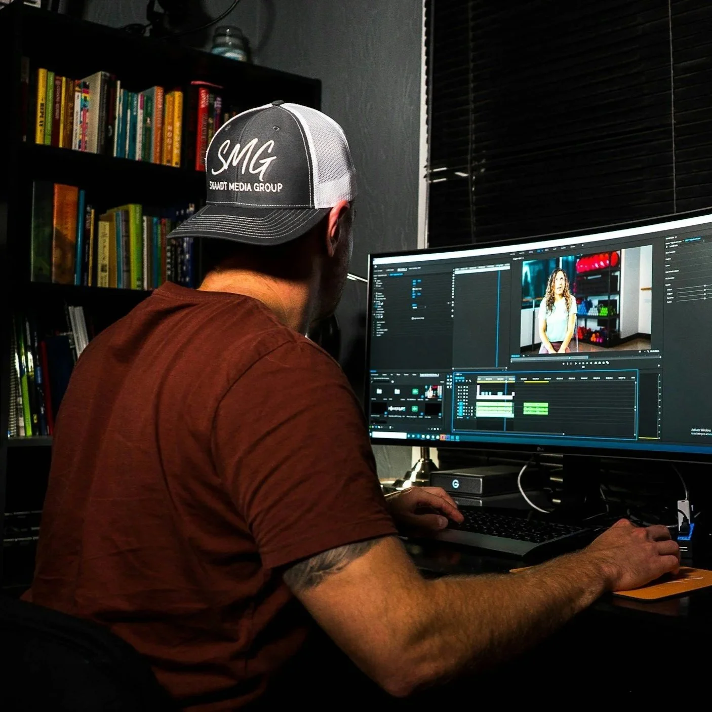 Man in a brown shirt and a black and white cap working on video editing on a computer at his desk, with a bookshelf in the background and a window with closed blinds.