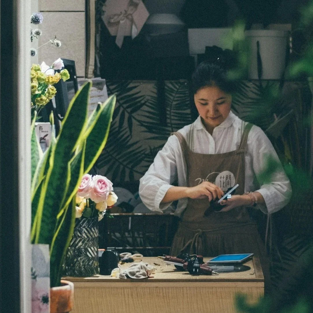 A woman looking at her phone inside a flower shop, with flowers and gardening tools on the table.