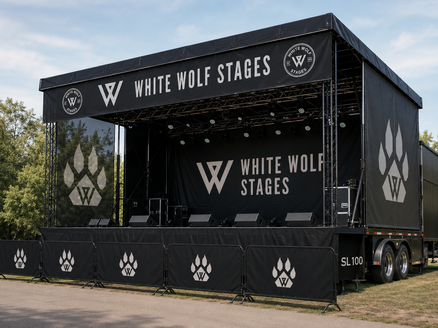 Empty outdoor stage with black banners and signage for 'White Wolf Stages,' featuring a wolf paw print logo, set up on a trailer with trees and a blue sky in the background.