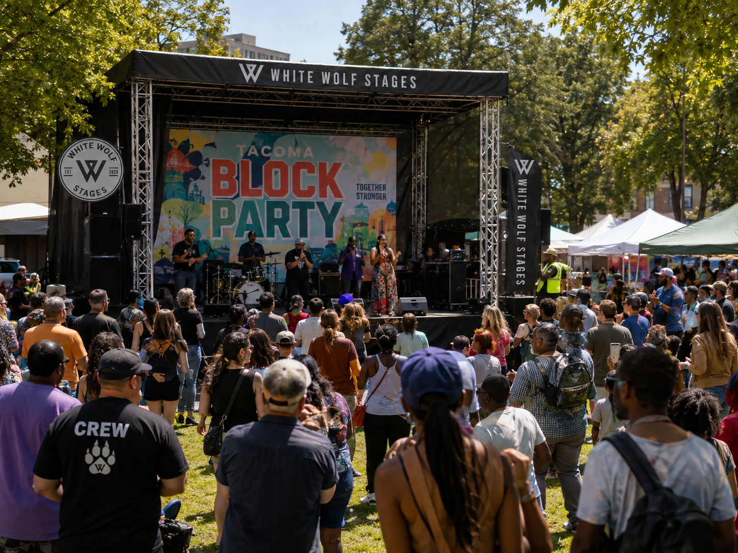 Crowd gathered outdoors watching a stage performance at the Tacoma Block Party with a large banner reading "White Wolf Stages" and a band playing on stage surrounded by trees and tents.