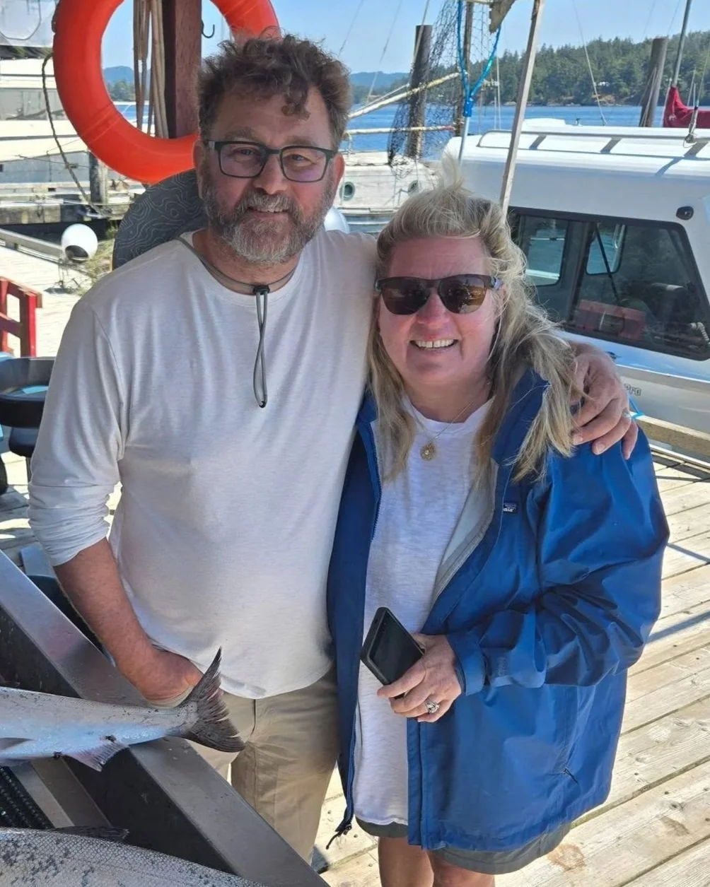 A smiling man and woman posing together at a marina with boats and docks in the background. The man is wearing a white long-sleeve shirt and glasses, the woman is wearing sunglasses and a blue jacket, holding a smartphone.