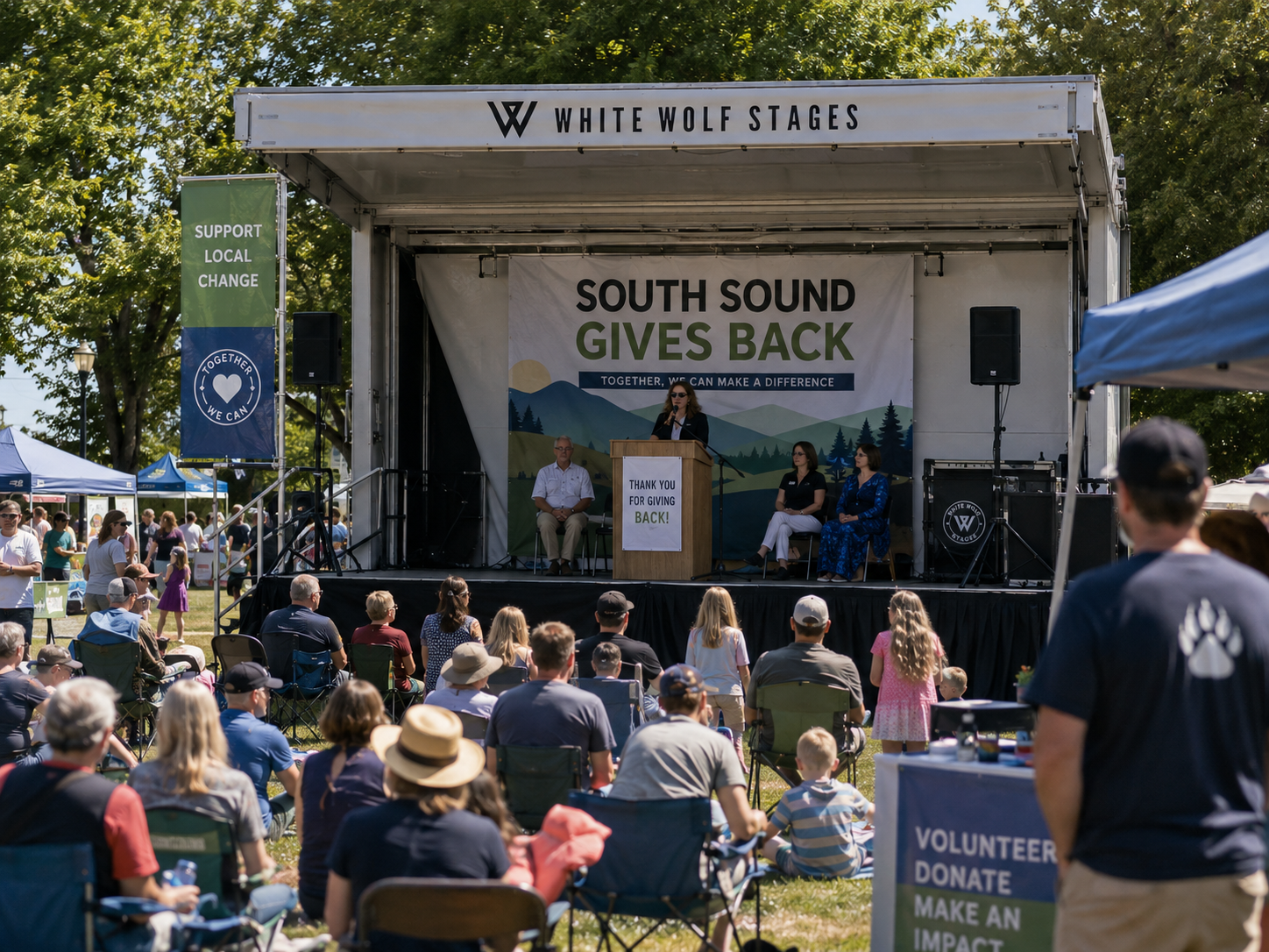 Outdoor community event with a stage labeled 'White Wolf Stages' featuring a large banner saying 'South Sound Gives Back.' An audience of diverse people, including children, watches a woman speaking at a podium; two people seated on stage. Tents and flags around the area hold messages supporting local change with a crowd enjoying a sunny day.