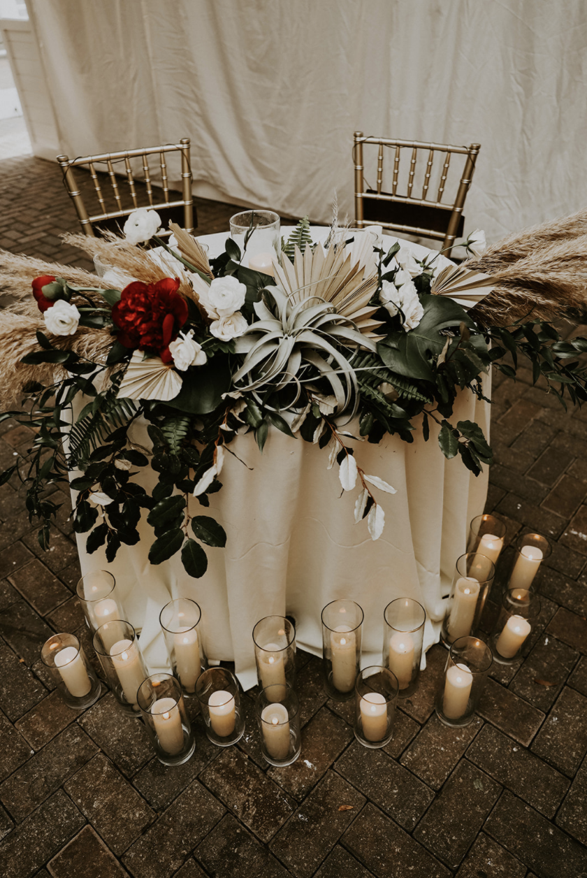 A round table with a beige tablecloth, decorated with an arrangement of white, red, and green foliage, surrounded by tall white candles in glass holders on a brick floor.