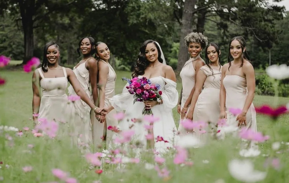 A group of seven women dressed in cream-colored dresses, standing in a grassy park with trees and pink flowers, one woman in the center holding a bouquet, suggesting a wedding celebration.