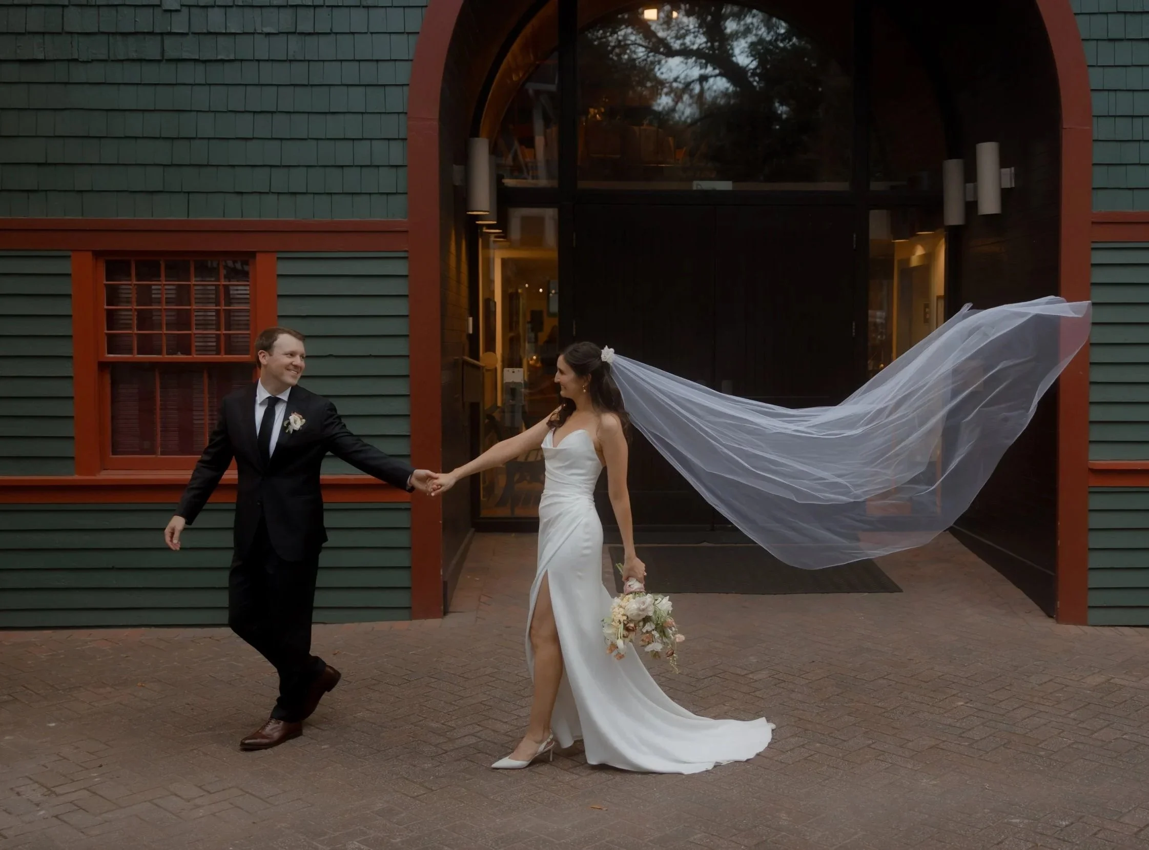 A newlywed couple holding hands and smiling outside a building, the bride in a white wedding gown with a flowing veil and the groom in a black suit with a white shirt and black tie.