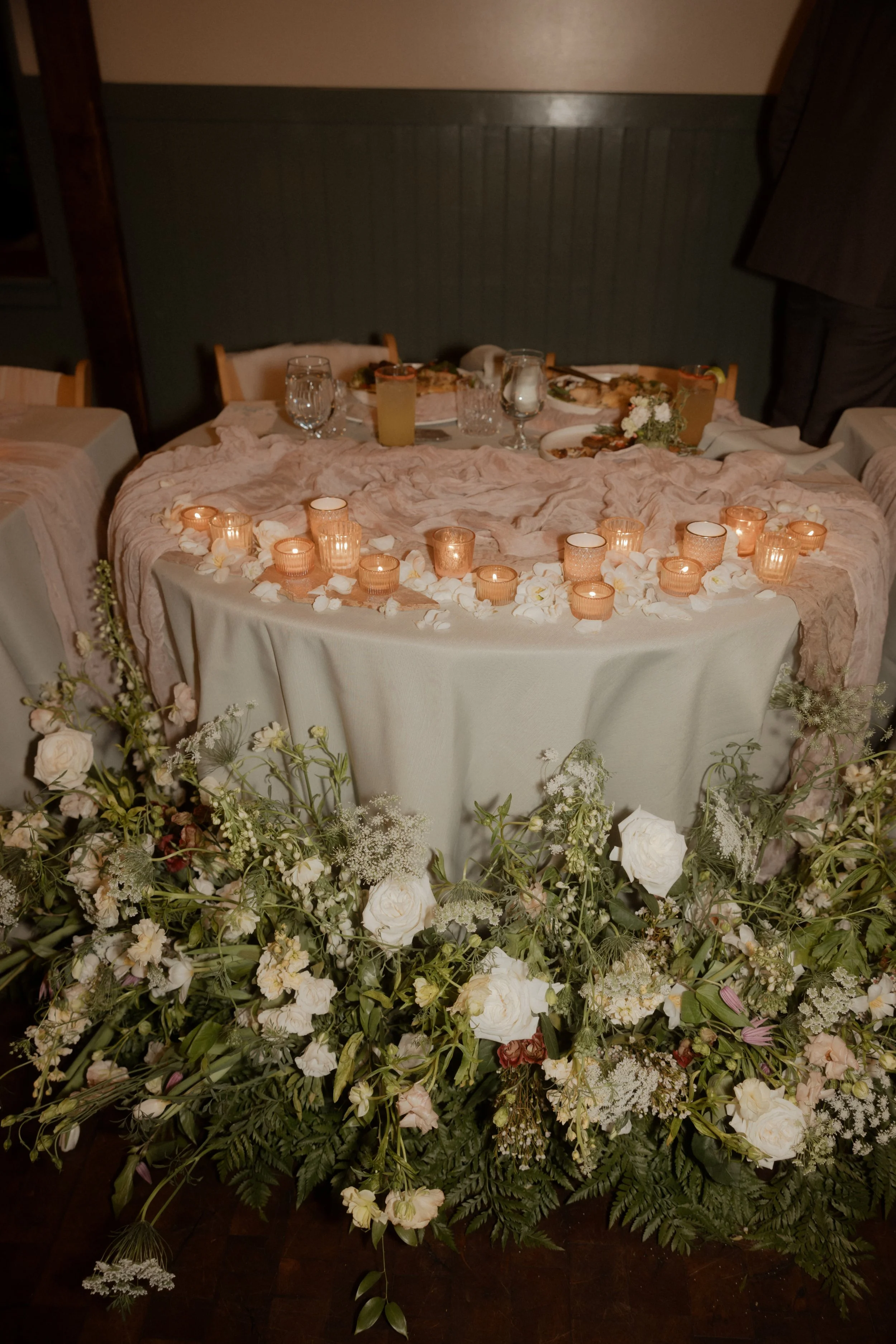 A wedding or event table decorated with flowers, candles, and glasses, surrounded by a floral arrangement on the floor.