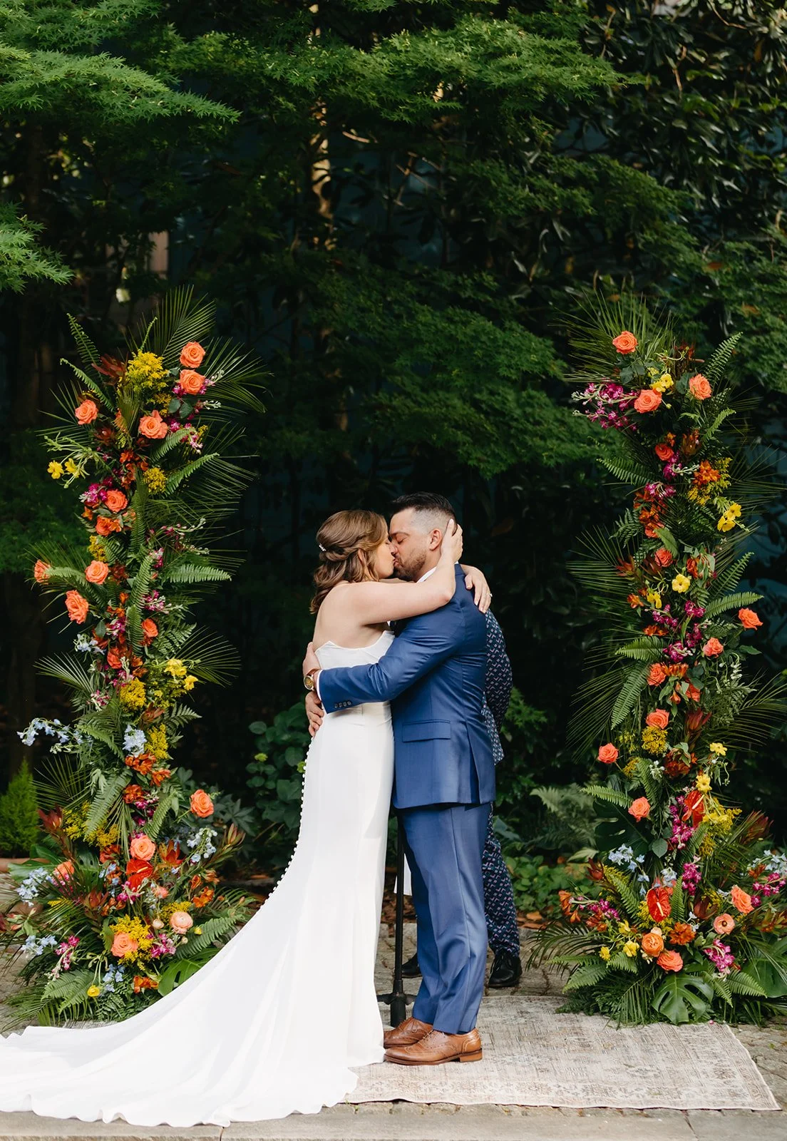 A couple in wedding attire kissing under a floral arch outdoors surrounded by green trees.