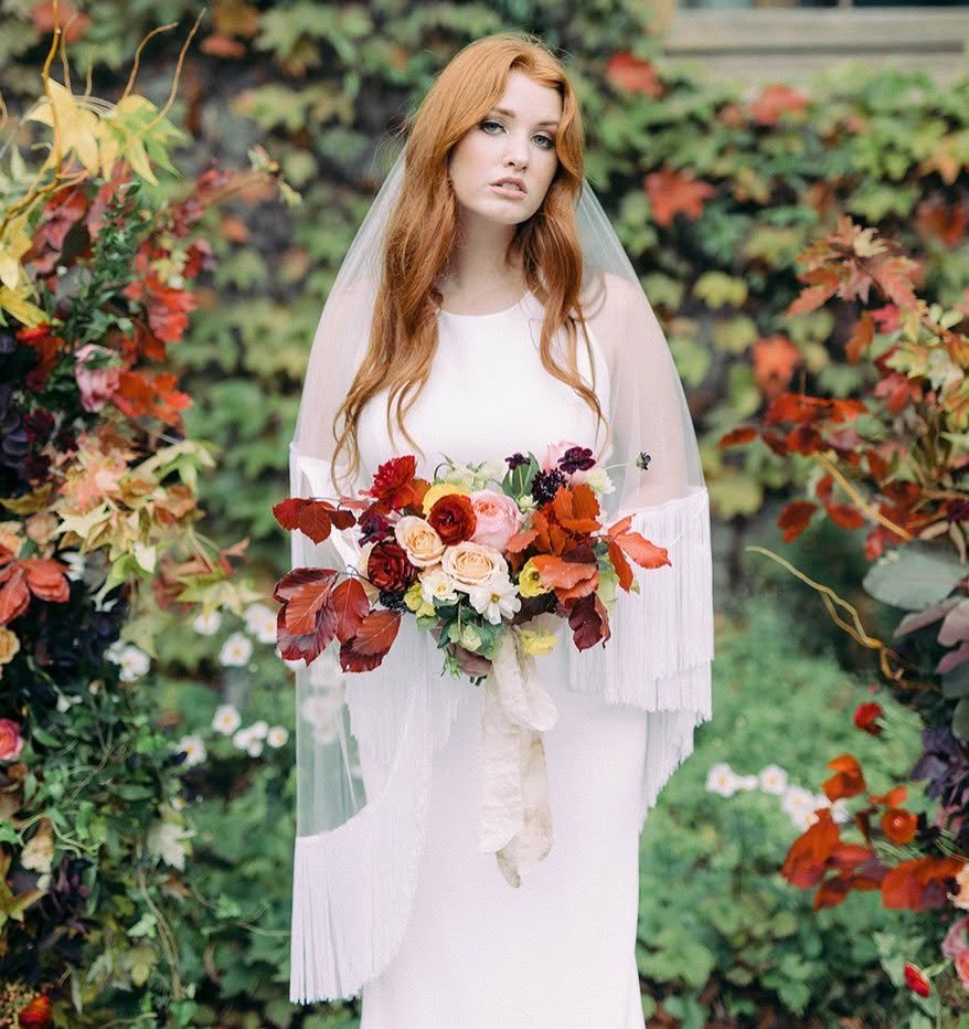 A young woman with long red hair in a white bridal gown and veil holding a bouquet of mixed flowers including roses, dahlias, and autumn leaves while standing outdoors surrounded by colorful foliage.
