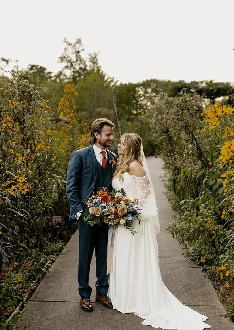 A bride and groom standing on a garden pathway, facing each other, surrounded by yellow and green foliage, with the bride holding a large bouquet of colorful flowers.