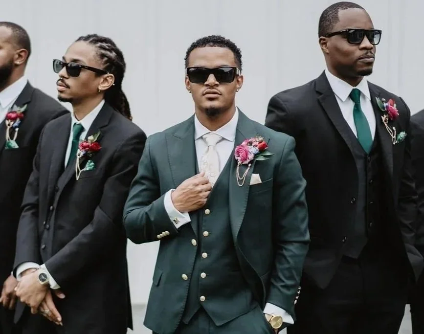 Four men dressed in formal suits and sunglasses standing outdoors, with boutonnieres on their lapels, during a ceremony.