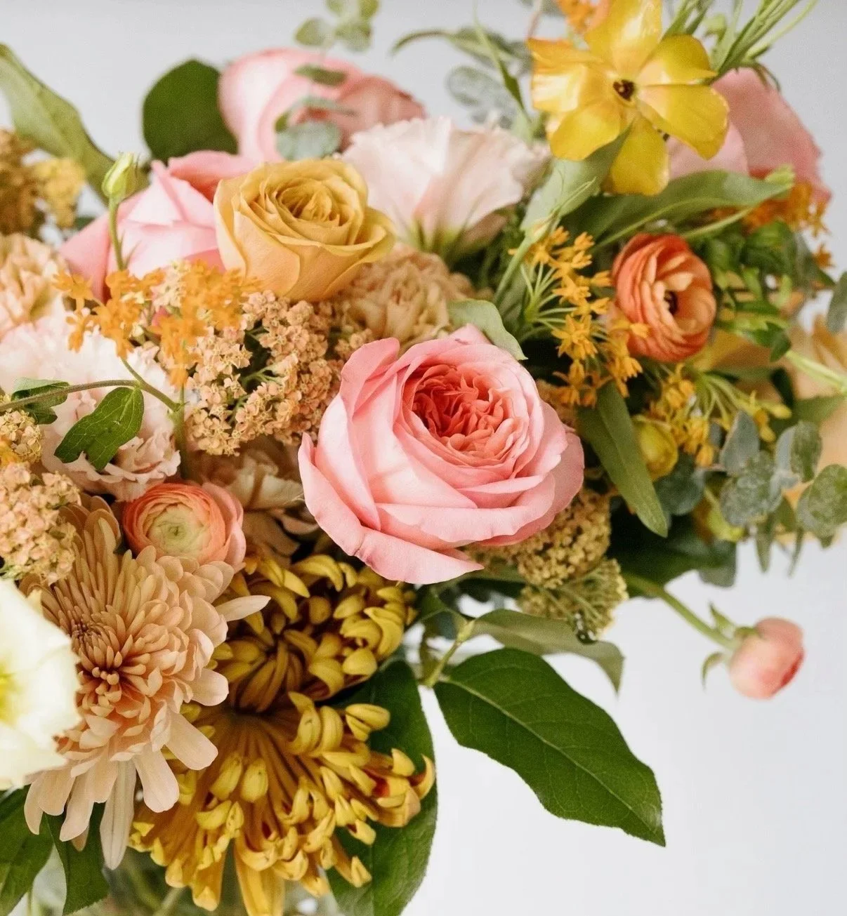 Close-up of a bouquet of mixed flowers including pink roses, yellow lilies, orange ranunculus, and chrysanthemums, with green leaves.