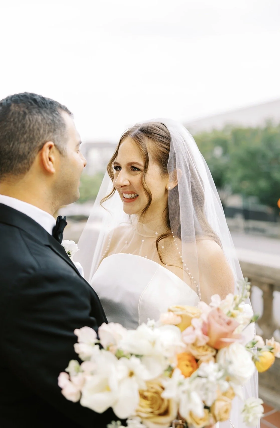A bride and groom stand close together outdoors, smiling at each other, with the bride holding a bouquet of flowers.