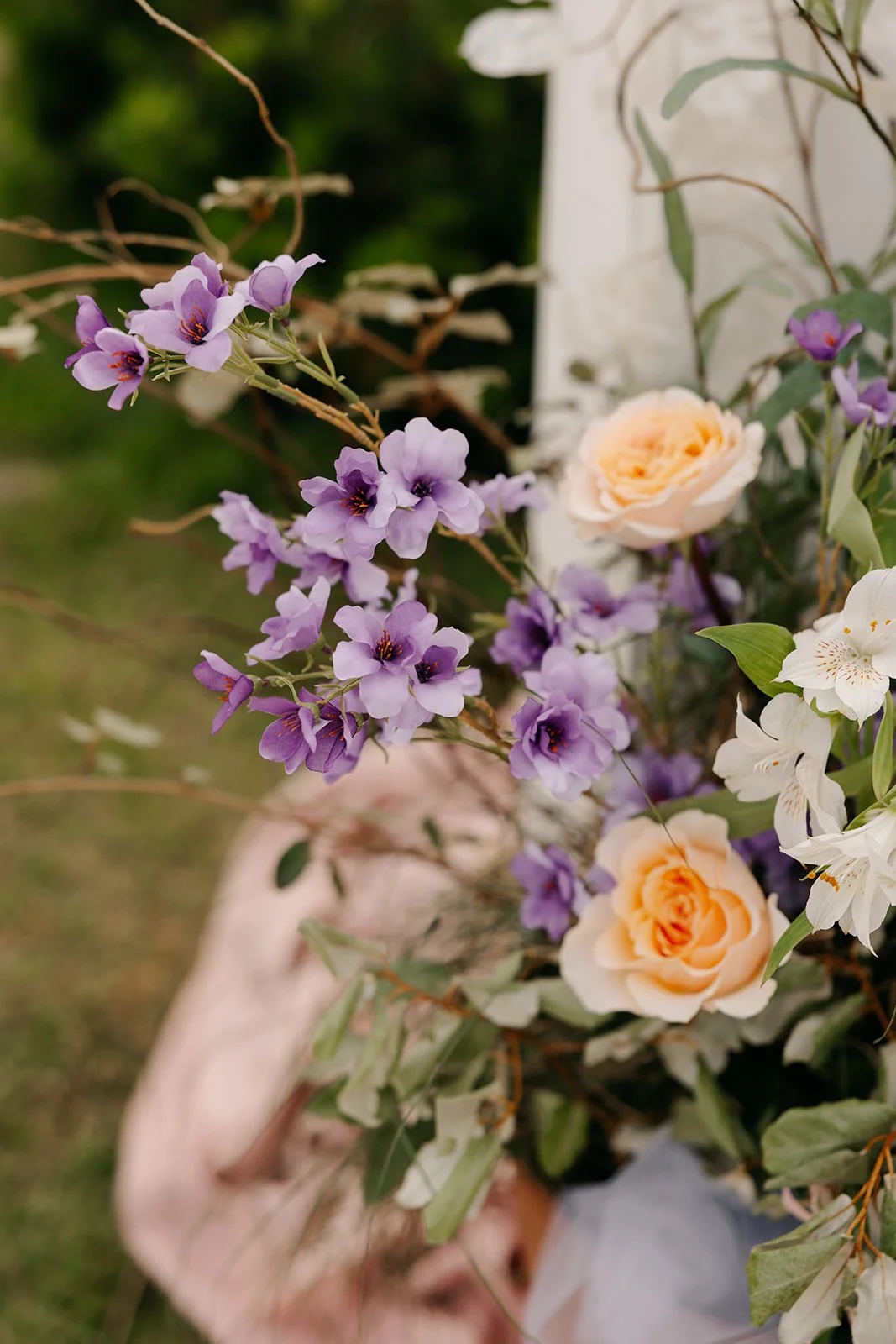 A bouquet of purple, peach, and white flowers with greenery, held by a person outdoors.