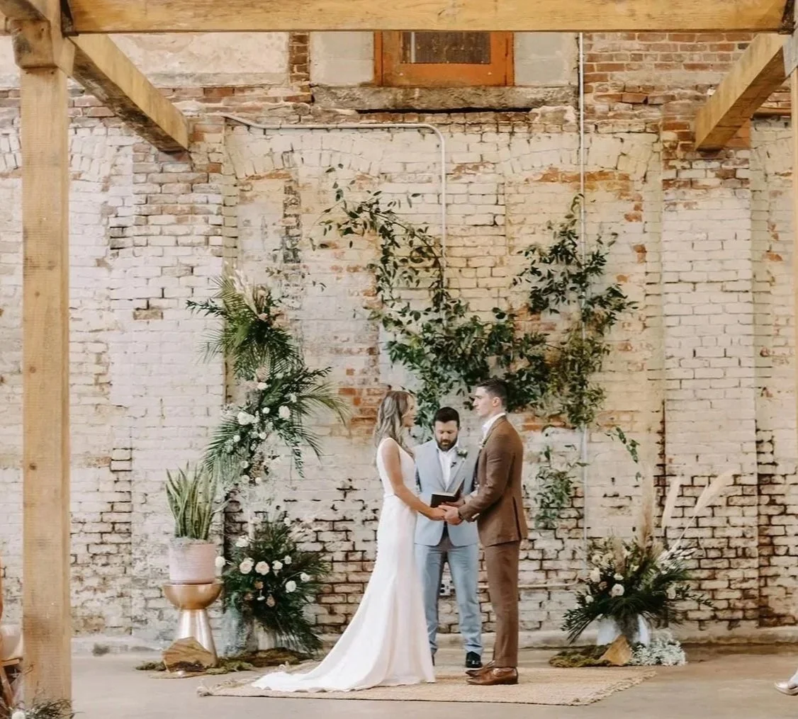 A couple getting married, holding hands and facing each other with an officiant in between, in a rustic indoor setting with exposed brick walls and greenery decorations.