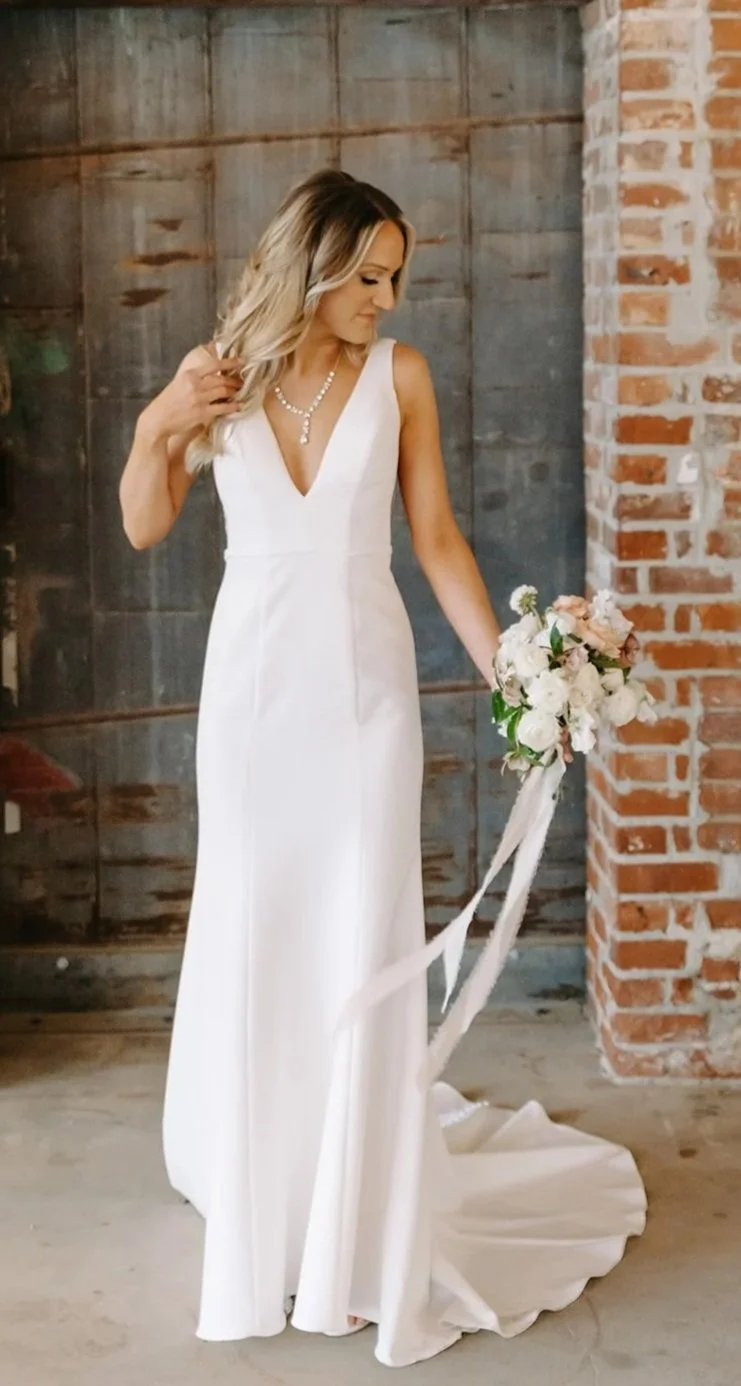 A woman in a white wedding dress holding a bouquet of flowers, standing indoors against a rustic brick and wooden wall.