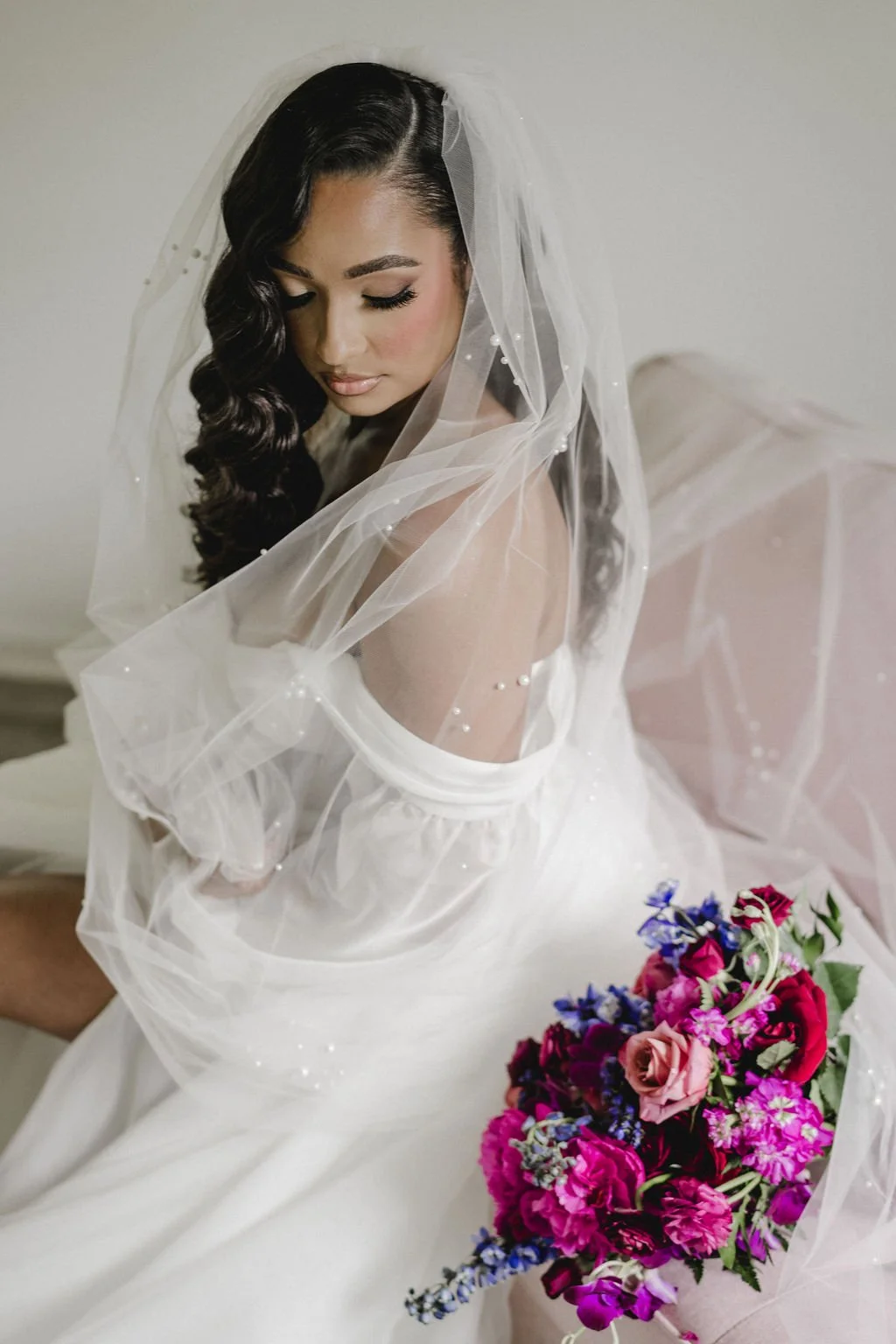 A bride with dark, wavy hair and makeup, wearing a white wedding gown and veil, holding a colorful bouquet of pink, purple, and blue flowers.