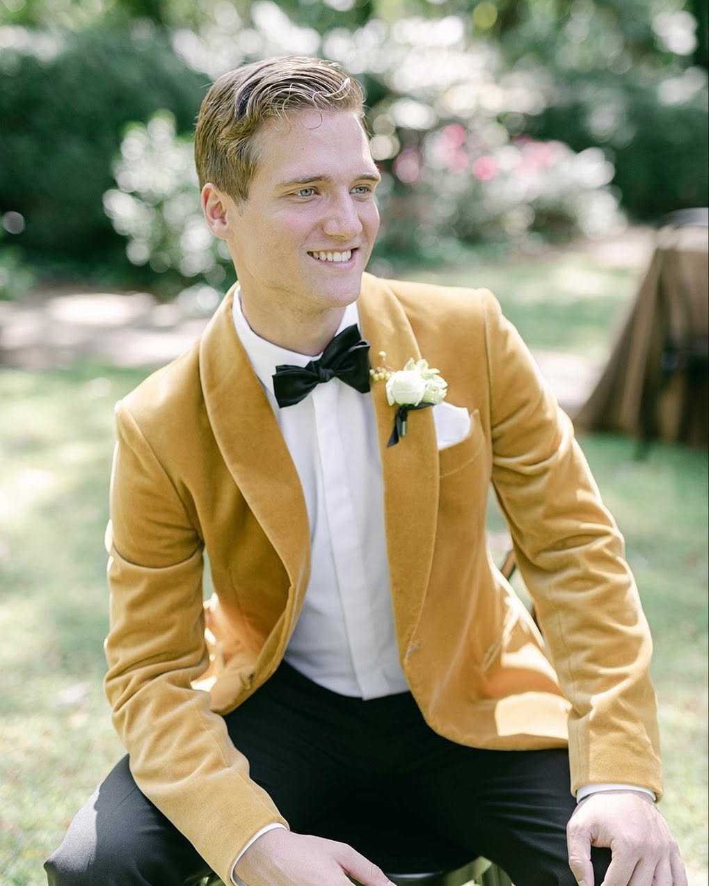 A young man in a mustard-colored velvet blazer, white dress shirt, black bow tie, and a white flower boutonniere, sitting outdoors on grass with a blurred background of trees and pink blossoms, smiling.