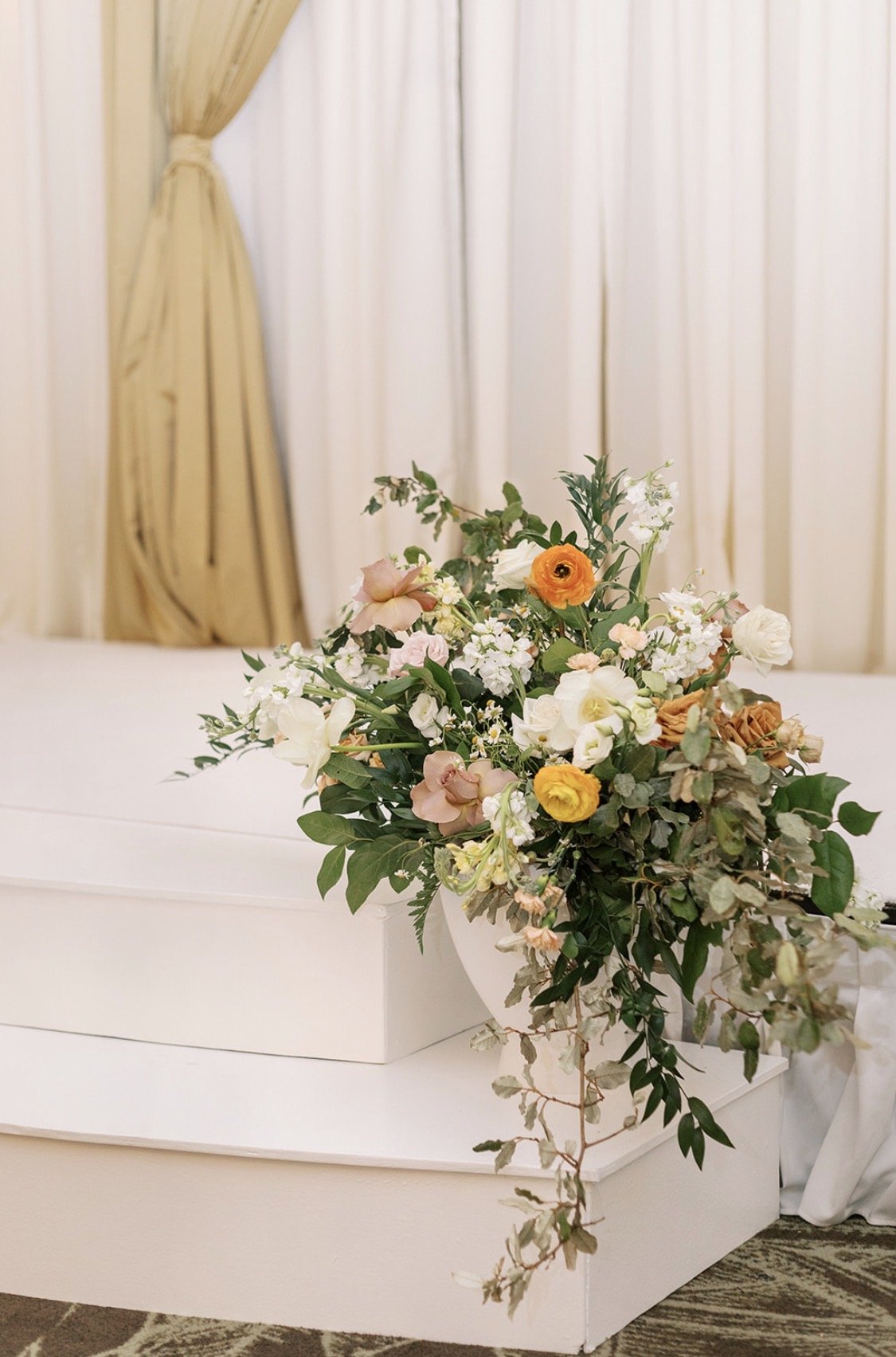 A floral arrangement with pink, white, yellow, and orange flowers and green foliage placed on a white surface with cream curtains in the background.