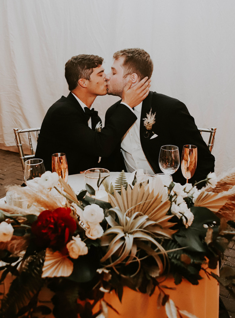 Two men in tuxedos sharing a kiss at a wedding reception, seated at a decorated table with floral arrangements and glassware.