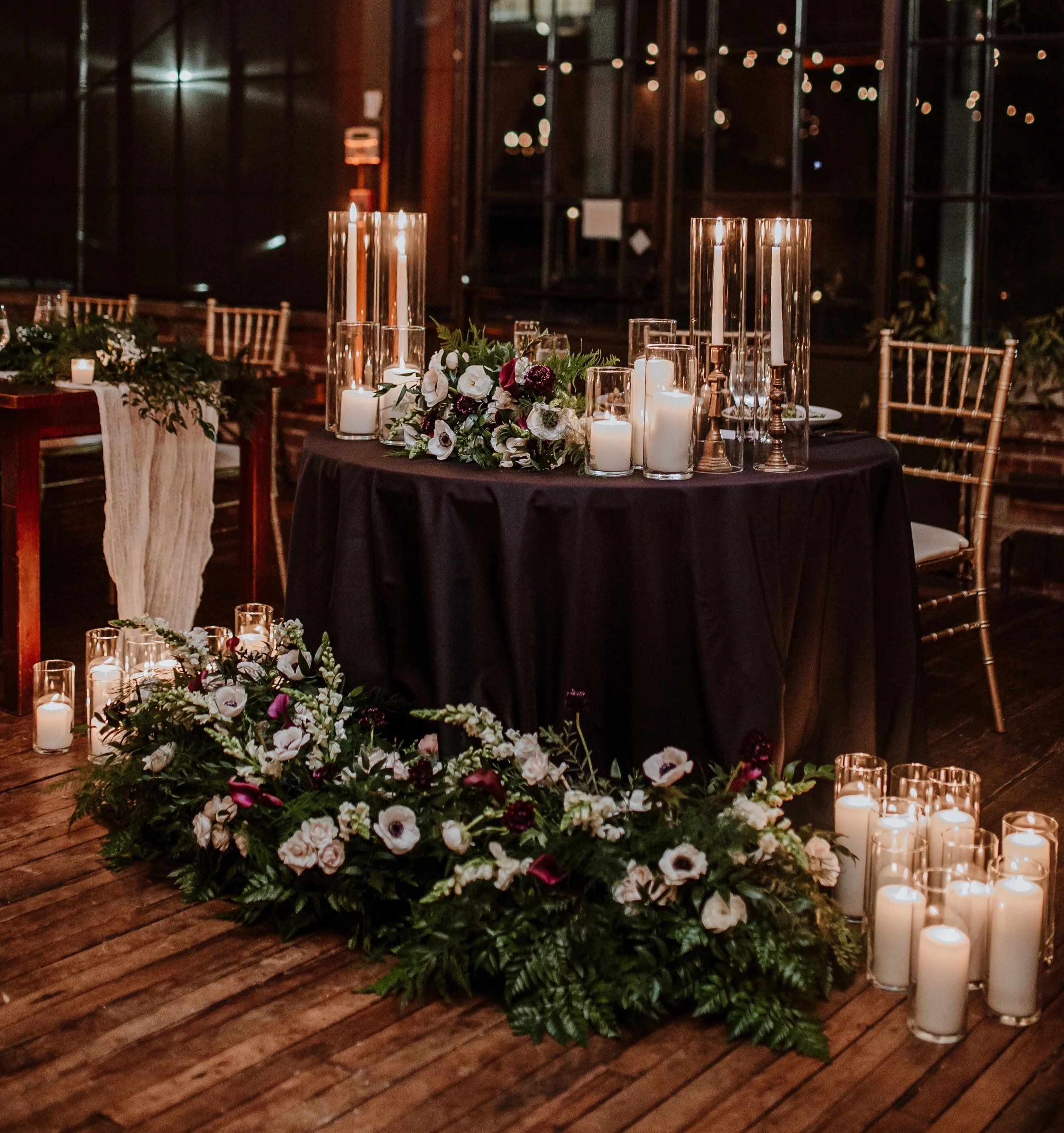 Elegant table centerpiece with white and dark purple flowers, tall white candles in glass holders, and gold-colored candlesticks, set on a black tablecloth in a dimly lit indoor venue with string lights outside the windows.
