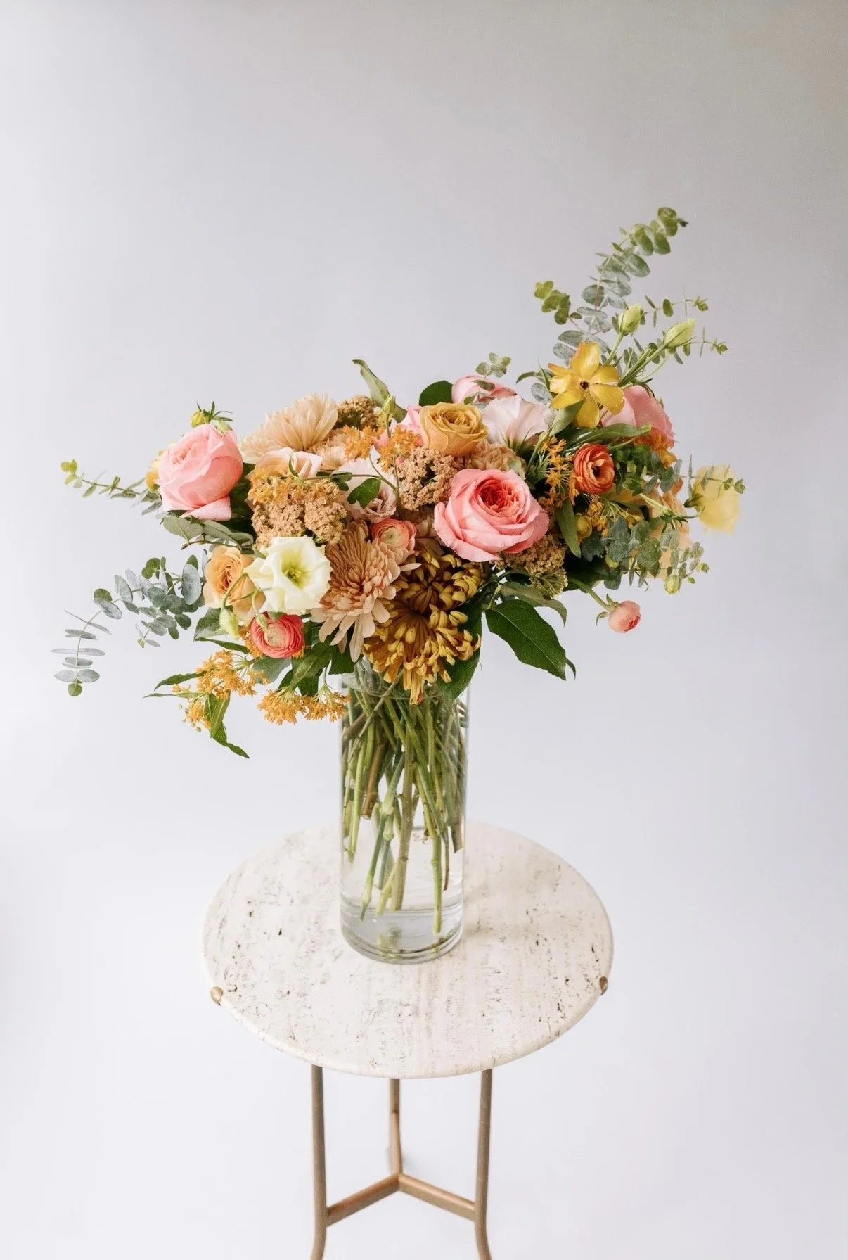 A glass vase filled with a mixed bouquet of pink, yellow, white, and orange flowers, with green foliage, placed on a round white table.