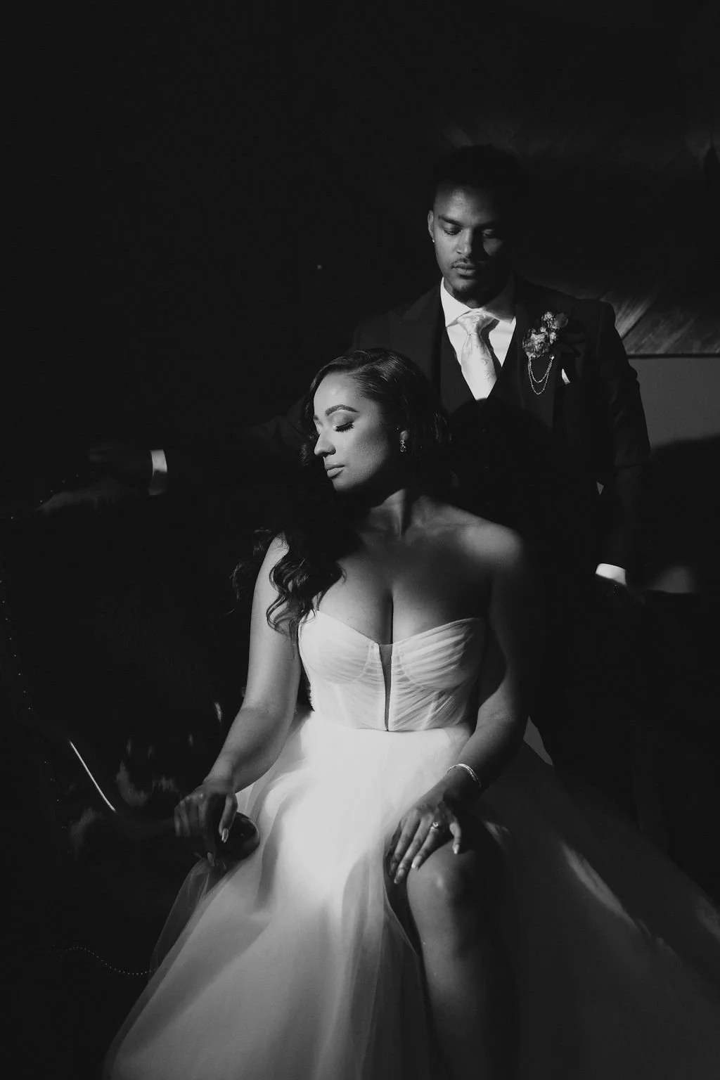 A black and white photo of a man in a suit and a woman in a wedding dress, with the woman sitting and the man standing behind her in a dimly lit setting.