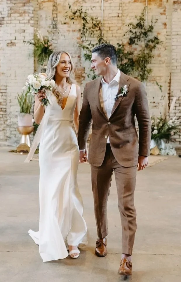 A bride and groom walking hand in hand in a rustic wedding venue, smiling at each other, with a brick wall and greenery in the background.