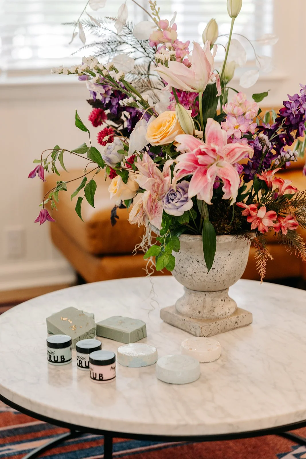 A white marble table with a large stone vase filled with a colorful flower arrangement, and small jars and soap bars in front of it.