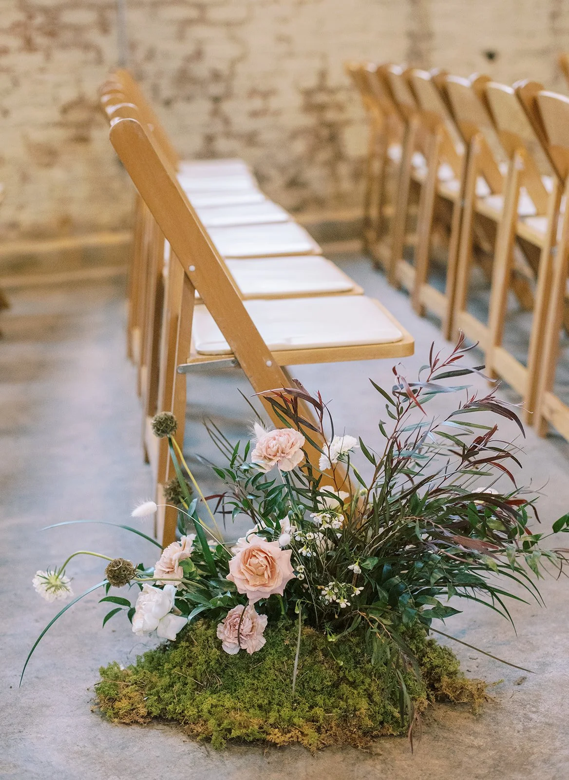 A flower arrangement with roses, greenery, and moss in front of wooden chairs with white cushions against a brick wall, possibly in a ceremony setting.