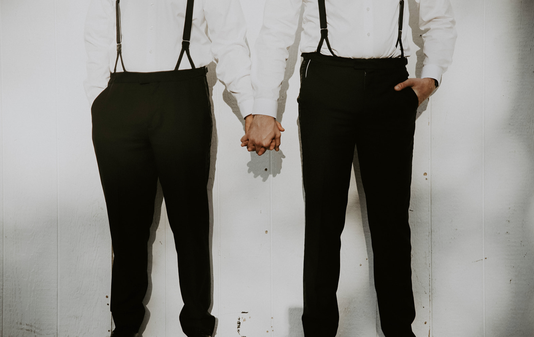 Two men dressed in white shirts and black pants holding hands against a white wall.