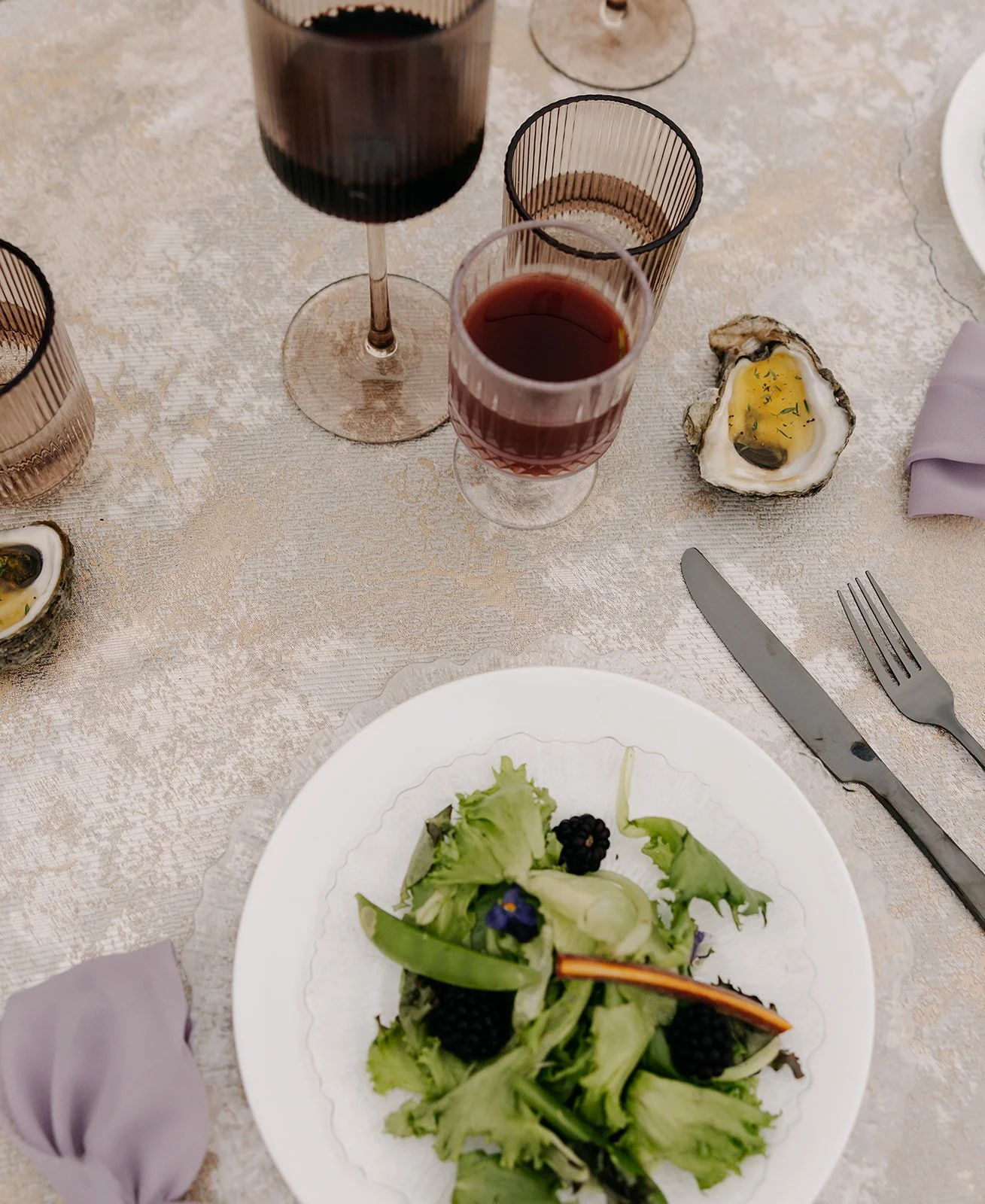 Table setting with a salad on a white plate, glasses of red and dark beverages, oysters, and a fork and knife on a textured tablecloth.