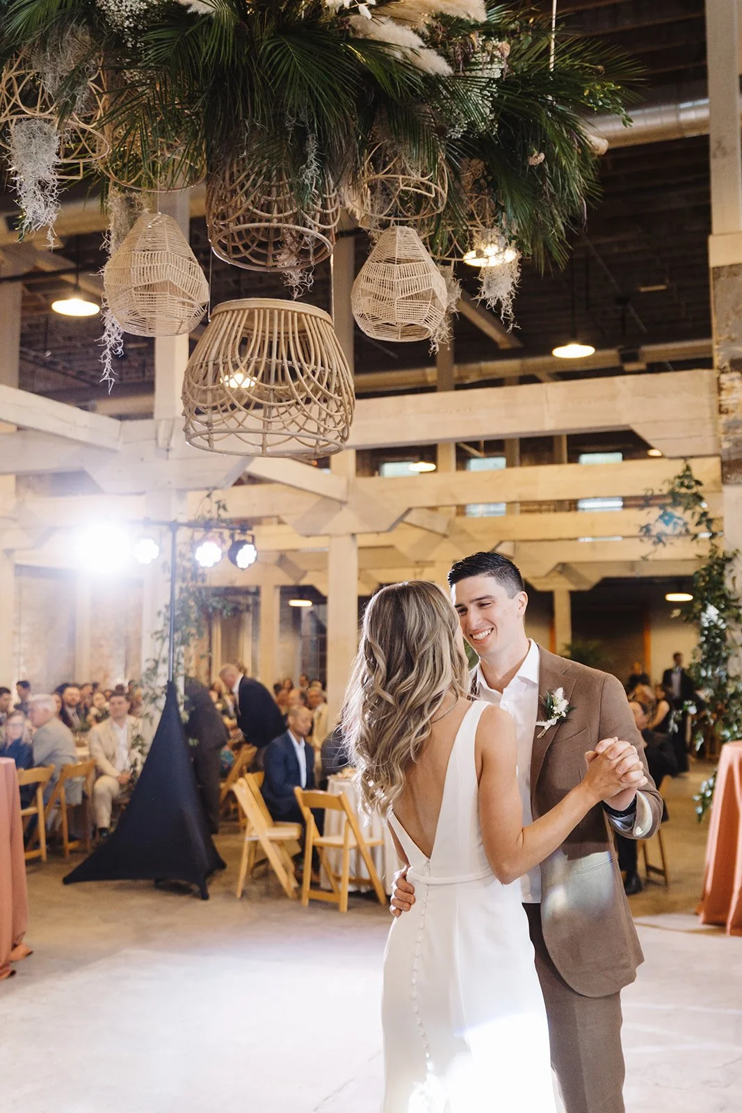 A bride and groom dancing at their wedding reception with guests seated at tables in the background, decorated with hanging wicker lanterns and lush greenery.