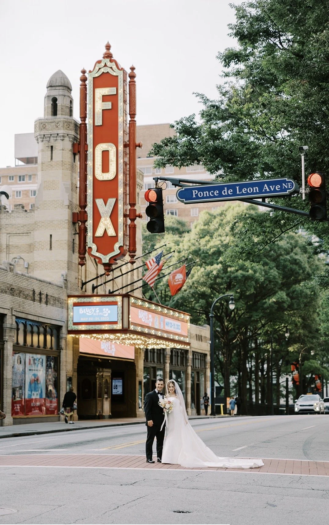 A bride and groom standing on a street in front of the Fox Theatre, with signs indicating the street is Ponce de Leon Avenue NE, and a marquee with lights. The bride is wearing a long white wedding dress and holding a bouquet, and the groom is dressed in a black suit.