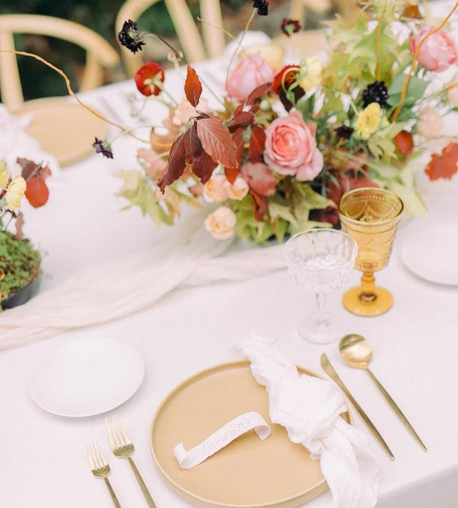 Elegant table setting with a large floral centerpiece, gold goblet, crystal glass, gold spoon, and fork on a white tablecloth, prepared for a special occasion or event.