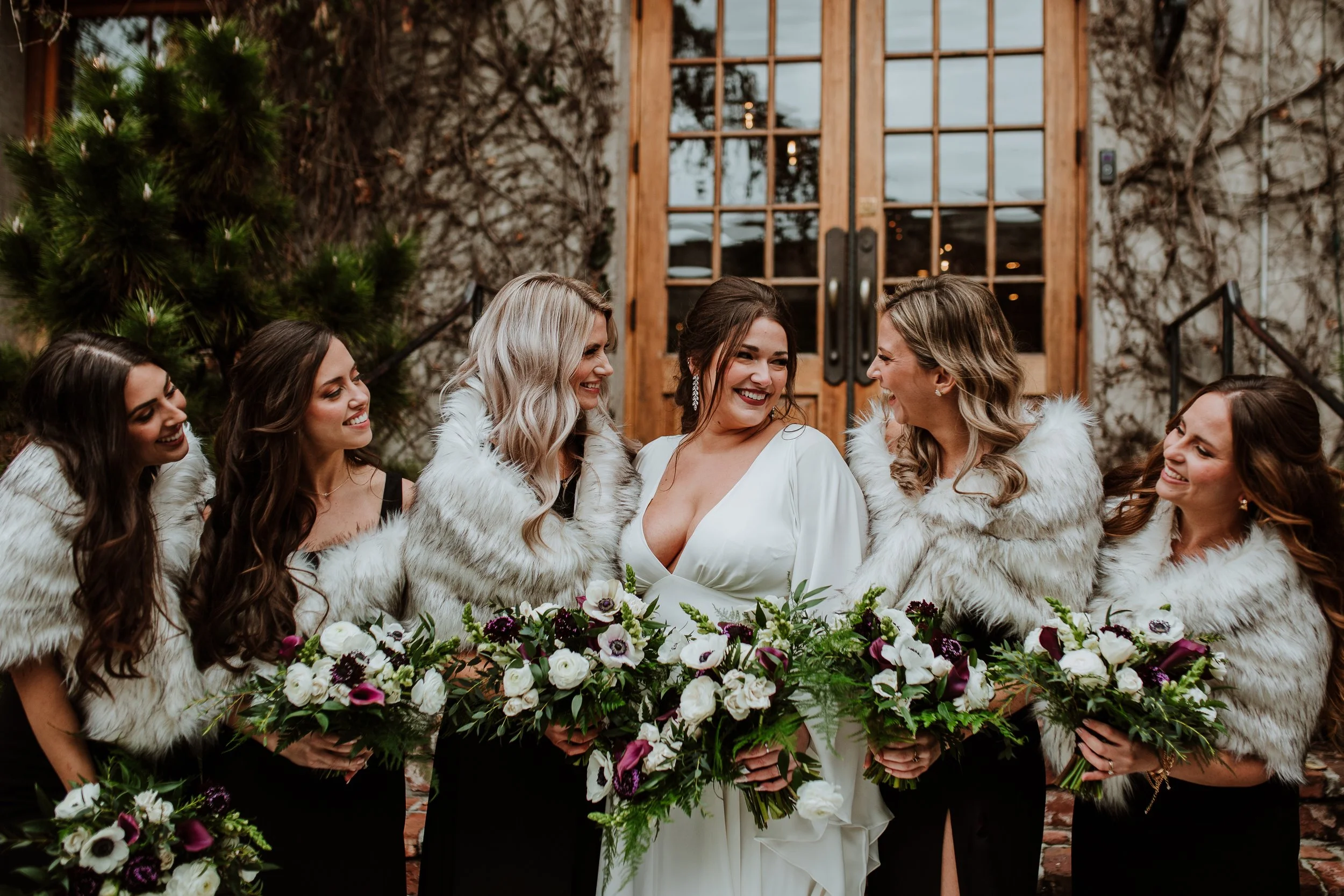 A group of women dressed in elegant attire with faux fur shawls, gathered outdoors in front of a rustic wooden door surrounded by climbing vines and trees. They are smiling and holding bouquets of flowers, seemingly celebrating a wedding.