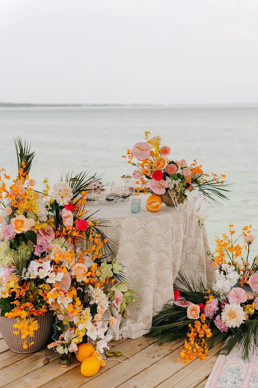 A decorated table with floral arrangements by a body of water, featuring pink, orange, and white flowers, with some citrus fruits nearby.