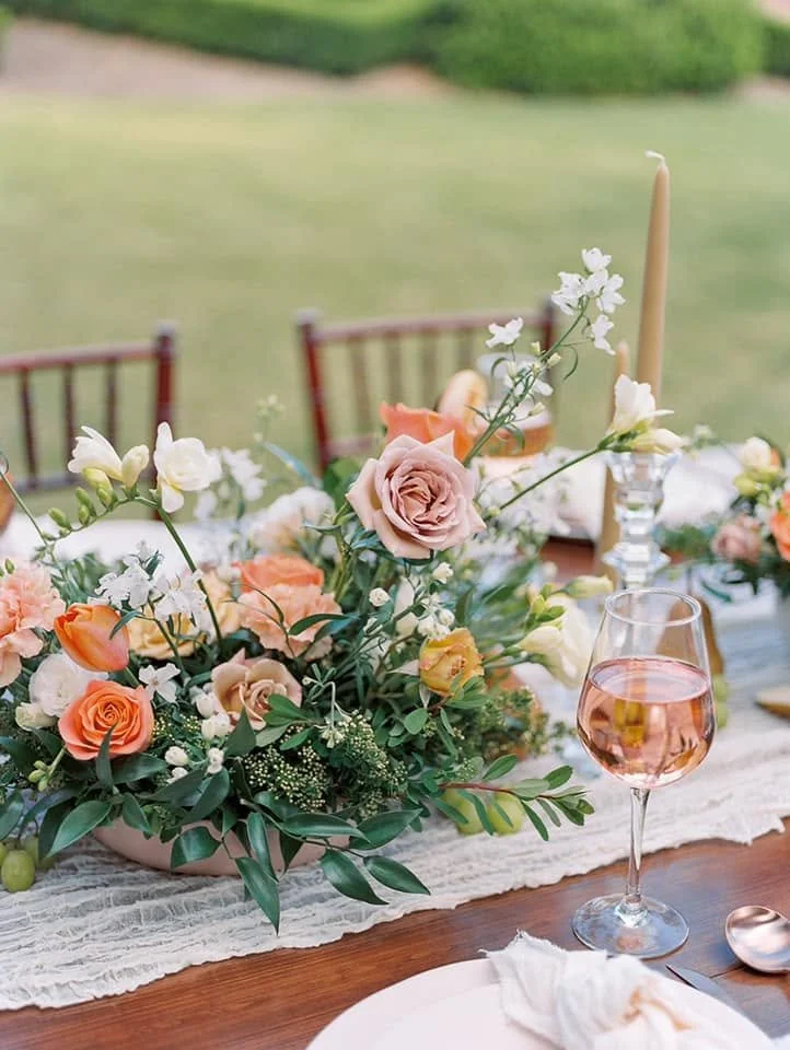 Elegant outdoor table setting with a floral centerpiece, a glass of pink wine, tall candlesticks, and chairs in the background on a grassy area.