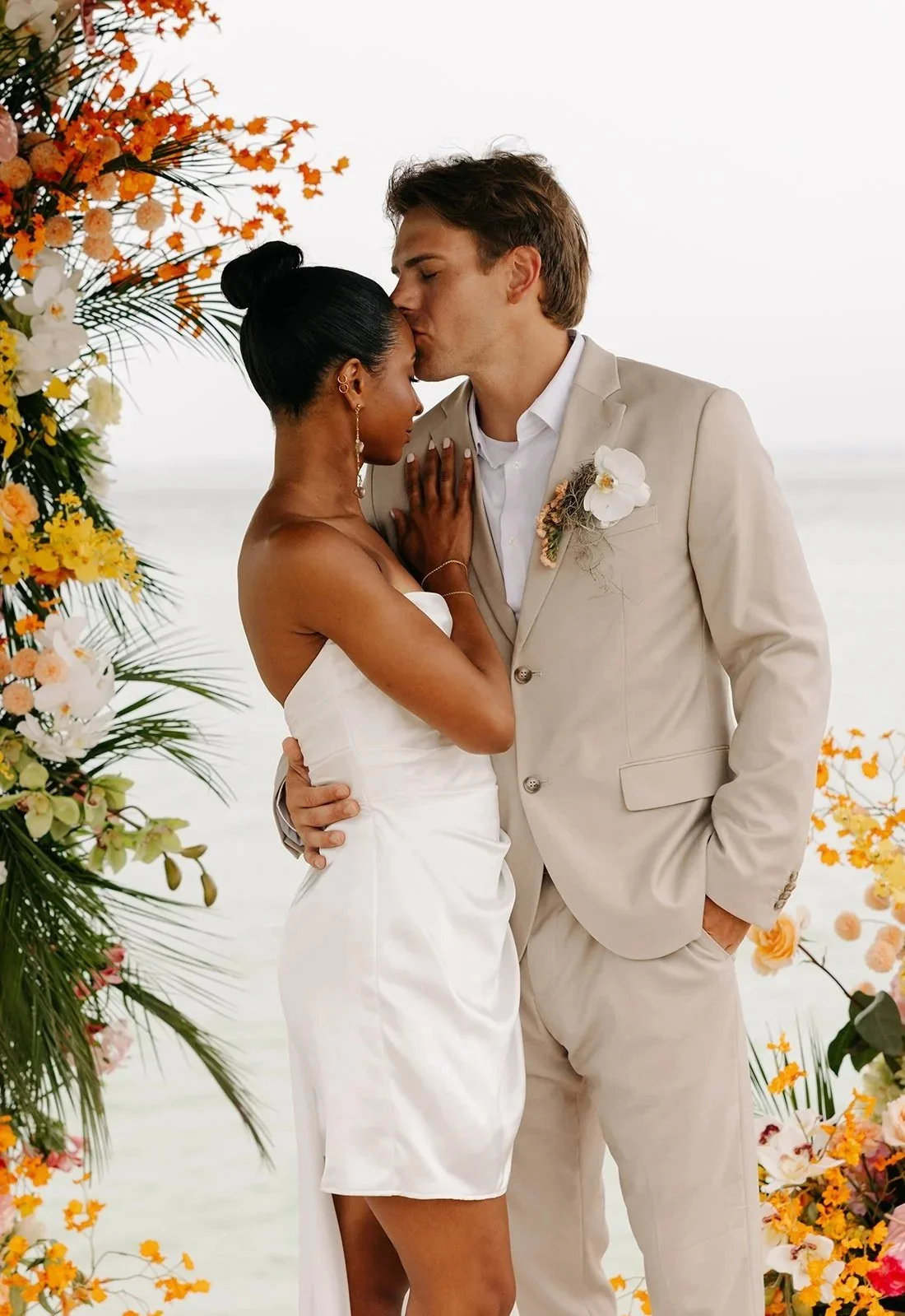A bride and groom share a kiss at their beach wedding, surrounded by colorful flowers and standing near the water.